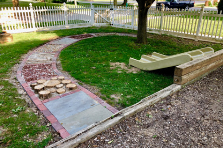 a path made of several materials, including wood, stones, and concrete. It is surrounded by grass and a single tree.