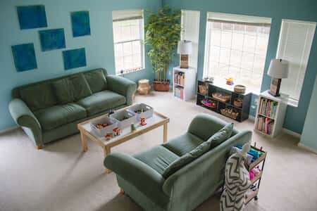 overhead view of a living room learning space with two couches facing each other, two small bookshelves with lamps atop, a low shelf with play materials between those, and a dress form with baskets of fabric behind the nearest couch.