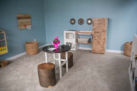 a home play space with tree stump seating on either side of a small table. Baskets and a play kitchen are in the background.