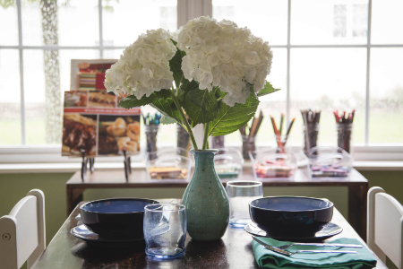 large flowers on vase in the middle of a small table. The table is set as if for a meal. A shelf with art matrials in the background.
