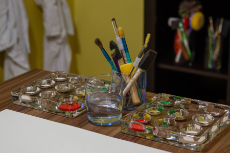 close up of paintbrushes in a glass cup on a wooden table. Another glass of water is in front of them. Glass paint palettes sit on either side.